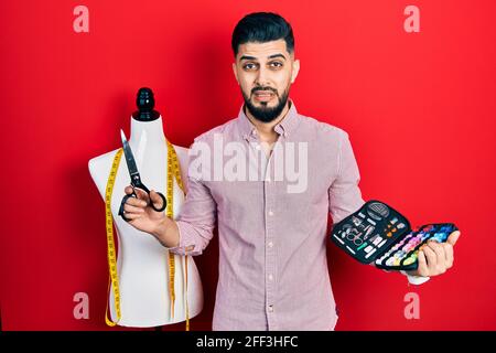 Schöner Schneider Mann mit Bart hält Näh-Kit und Schere ahnungslos und verwirrt Ausdruck. Zweifel Konzept. Stockfoto
