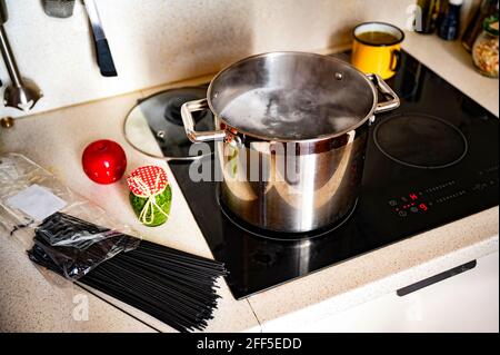 Kochendes Wasser im Topf auf heissem Keramikkochfeld, Beutel mit schwarzen Spaghetti, Flasche mit grünem Basilikum-Pesto, Küchenuhr für roten Apfel, kleiner gelber Topf mit Suppe. Stockfoto