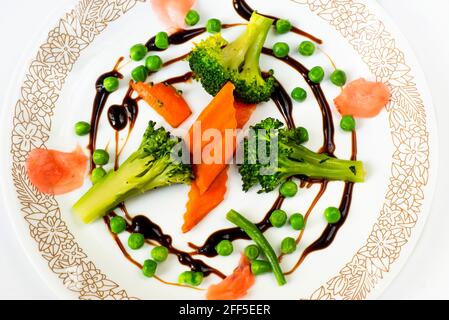 Gesunde und schön arrangierte pflanzliche Nahrung - geschmorte Karotten, Brocoli, Erbsen, eingelegter Ingwer in kreisrunder Anordnung mit Balsamico auf weißem Teller. Stockfoto