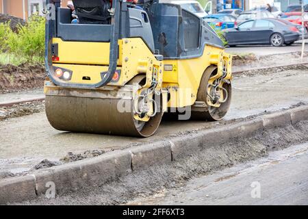 Eine gelbe Straßenwalze, die das Fundament eines Bürgersteiges auf einer Baustelle entlang einer Betoneinfassung in der Nähe der Fahrbahn stampft. Speicherplatz kopieren. Stockfoto