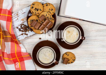 Zwei Tassen Kaffee mit Schokoladenkeksen und Schokolade auf dem Hintergrund eines offenen Buches. Heißer Kaffee mit Schokoladenkeksen im Tee Stockfoto