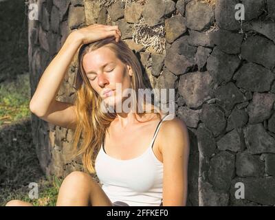 Ein junges Mädchen sitzt in der Abendsonne an einer alten Steinmauer gelehnt. Sie hat ihre Augen geschlossen und ihr rechter Arm befindet sich über ihrem Kopf. Sie hat Long blon Stockfoto