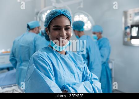 Lächelnde Mischchirurin mit Gesichtsmaske und Schutzfunktion Kleidung im Operationssaal Stockfoto