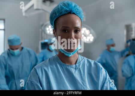 Lächelnde Mischchirurin mit Gesichtsmaske und Schutzfunktion Kleidung im Operationssaal Stockfoto