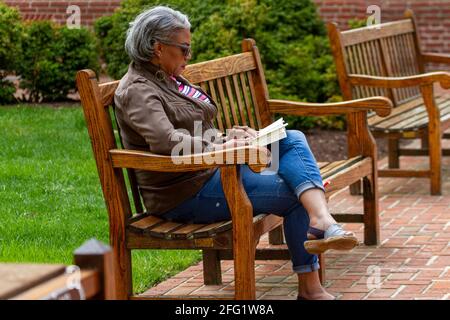 Easton, MD, USA 04-18-2021: Eine ältere afroamerikanische Frau sitzt allein auf einer Holzbank im Park und liest ein Buch, das das schöne Wetter genießt Stockfoto