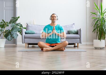 Yoga zu Hause üben. Workout zu Hause. Mann mittleren Alters mit weißen Kopfhörern meditiert während des Yoga. Gebetshaltung. Stockfoto