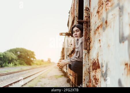 Porträt eines lächelnden, gutaussehenden asiatischen Mannes, der am öffentlichen Bahnhof steht. Junger Mann steht im alten Vintage-Zug Stockfoto