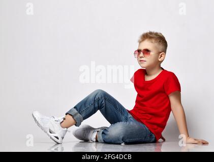 Der normale blonde Junge sitzt in rotem T-Shirt, blauer Jeans, weißen Turnschuhen und Sonnenbrille auf dem Boden und lehnt sich an seine Hände Stockfoto