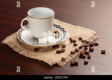 Porzellanbecher mit Untertasse und goldenem Löffel auf Jute-Serviette. Vintage-Kaffeeset auf Holztisch, mit Zucker und Kaffeebohnen Stockfoto
