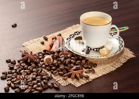 Kaffee in Porzellanbecher auf Untertasse mit Zimt, Muskatnuss und Sternanise auf der Serviette von aute. Vintage-Kaffeeset auf Holztisch mit abgeworfenen Kaffeebohnen. Stockfoto