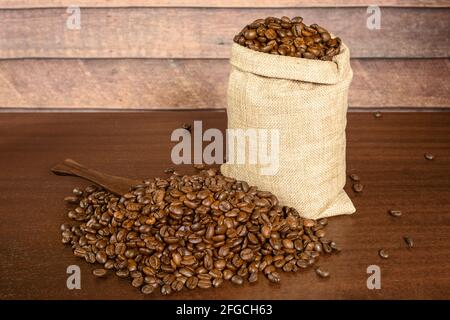 Kaffeebohnen. Geröstete Kaffeebohnen in Jute-Burlack-Sack und viel verstreuter Kaffee mit Spachtel, auf dem braunen Holztisch mit rustikalem Hintergrund. Stockfoto