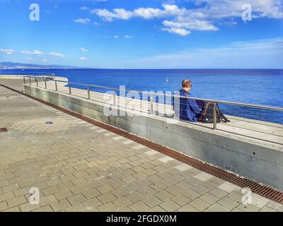Rückansicht eines älteren Mannes, der auf einer Promenade in der Nähe des Meeres sitzt. Stockfoto