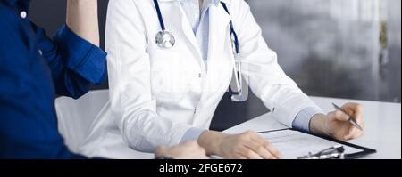Unbekannte junge Ärztin hört ihrer Patientin zu, während sie in der Klinik zusammen sitzt. Medizinkonzept während der Coronavirus-Pandemie Stockfoto