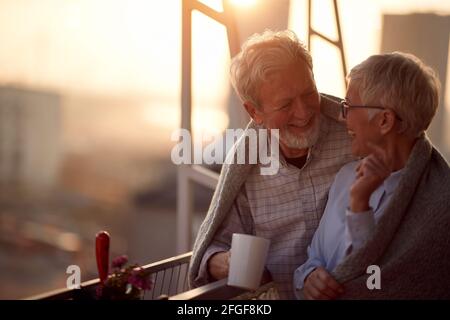 Ein älteres Paar, das bei einem wunderschönen Sonnenuntergang auf der Terrasse seiner Wohnung eine schöne Zeit miteinander hat. Ehepartner, Rentner, zusammen, zu Hause Stockfoto