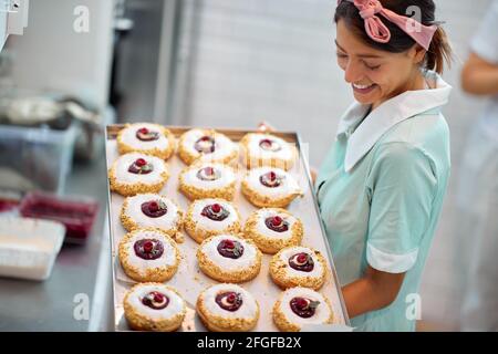 Ein schönes Retro aussehendes junges Mädchen in einer fröhlichen Atmosphäre In einer Süßwarenwerkstatt halten und auf ein Tablett schauen Mit leckeren Donuts bereit für eine Vergangenheit Stockfoto