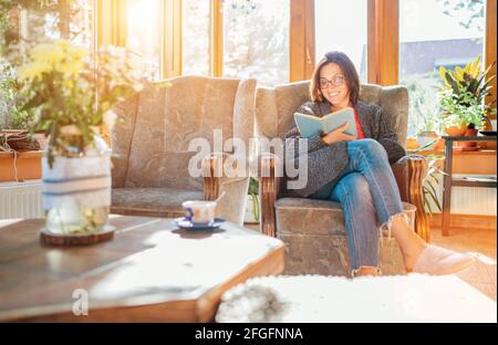 Fröhlich lächelnd weiblich gekleidet gemütliche Hauskleidung sitzen in bequemen Sessel, entspannen, ein Buch lesen und trinken Morgenkaffee oder Tee in Hous Stockfoto