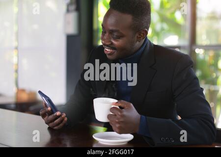 Afrikanischer Geschäftsmann trinkt einen heißen Kaffee im Café mit Blick neuigkeiten auf dem Smartphone Stockfoto