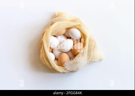 Hühnereier weiß und braun in einem beigefarbenen wiederverwendbaren Netzbeutel auf einem weißen Tisch. Konzept für landwirtschaftliche Produkte, natürliche Ernährung und Umweltschutz Stockfoto
