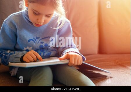 Ein süßes, kleines europäisches Mädchen im Alter von sieben Jahren liest ein Buch, bleibt zu Hause während der Selbstisolierung des Covid-19 Corona Virus Quarantäne. Flare Stockfoto