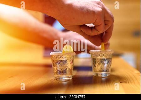 Zwei Gläser Tequila auf dem Hintergrund eines Laptops mit Blick auf einen Film. Männerhände legte eine Zitrone. Ein angenehmer Abend zu Hause. Warm gedämpfte Beleuchtung. fl Stockfoto