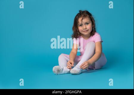 Schöne Kind Ballerina Mädchen binden Schnürsenkel auf Ballettschuhe sitzen Auf einem blauen Hintergrund und lächelnd niedlich Blick auf die Kamera Stockfoto