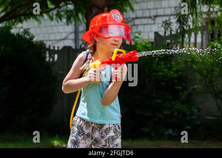 Feuerwehrmann, kleines Kind, kleines Mädchen im Schulalter, das einen Feuerwehrhelm trägt und mit einer Wasserpistole im Garten spielt. Träume, zukünftiger Beruf, Arbeit, Spiel Stockfoto