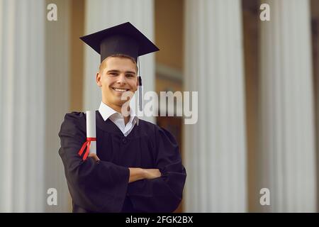 Erfolgreicher Absolvent im akademischen Kleid posiert mit Diplom in den Händen vor Hochschulhintergrund. Stockfoto