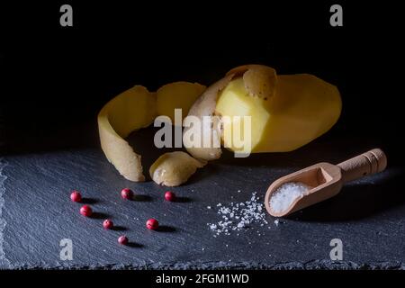 Eine geschälte Kartoffel, auf der die Haut aufliegt, rote Beeren und ein Holzlöffel voller Salzkörner auf einem Schieferteller Stockfoto