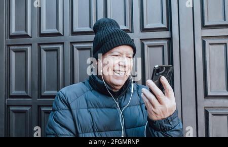 Lächelnder älterer Mann macht einen Videoanruf mit seinem Schwarzen Smartphone und weiße Kopfhörer im Garten zu Hause Stockfoto