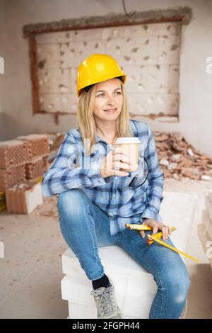Hübsche junge Arbeiterin mit gelbem Schutzhelm nimmt Kaffee Pause auf Baustelle innen und ist glücklich Stockfoto