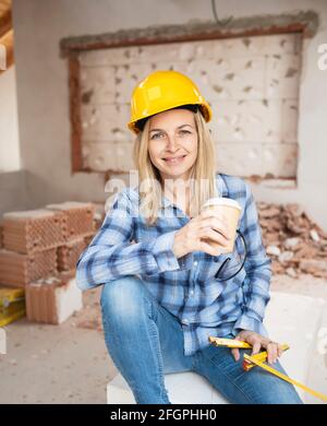 Hübsche junge Arbeiterin mit gelbem Schutzhelm nimmt Kaffee Pause auf Baustelle innen und ist glücklich Stockfoto