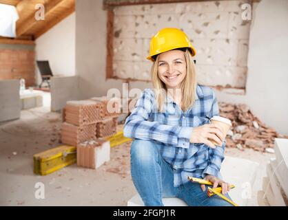 Hübsche junge Arbeiterin mit gelbem Schutzhelm nimmt Kaffee Pause auf Baustelle innen und ist glücklich Stockfoto