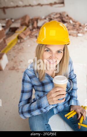Hübsche junge Arbeiterin mit gelbem Schutzhelm nimmt Kaffee Pause auf Baustelle innen und ist glücklich Stockfoto