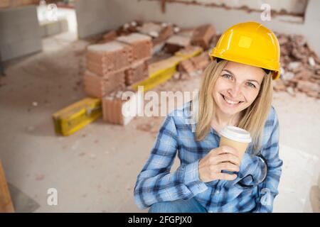 Hübsche junge Arbeiterin mit gelbem Schutzhelm nimmt Kaffee Pause auf Baustelle innen und ist glücklich Stockfoto