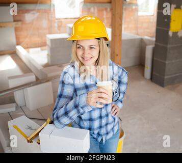 Hübsche junge Arbeiterin mit gelbem Schutzhelm nimmt Kaffee Pause auf Baustelle innen und ist glücklich Stockfoto