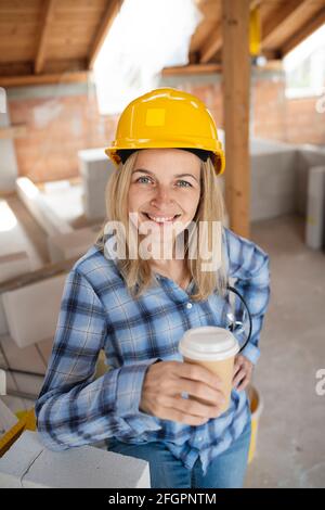 Hübsche junge Arbeiterin mit gelbem Schutzhelm nimmt Kaffee Pause auf Baustelle innen und ist glücklich Stockfoto