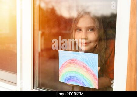 Laute Wirkung. Kind Mädchen sieben Jahre alt mit Zeichnung Regenbogen schaut durch das Fenster während covid-19 Quarantäne. Zu Hause bleiben, lassen Sie uns alle gut sein. Flare Stockfoto