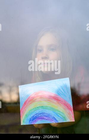 Laute Wirkung. Kind Mädchen sieben Jahre alt mit Zeichnung Regenbogen schaut durch das Fenster während covid-19 Quarantäne. Zu Hause bleiben, lassen Sie uns alle gut sein. verti Stockfoto