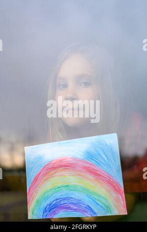 Laute Wirkung. Kind Mädchen sieben Jahre alt mit Zeichnung Regenbogen schaut durch das Fenster während covid-19 Quarantäne. Zu Hause bleiben, lassen Sie uns alle gut sein. verti Stockfoto