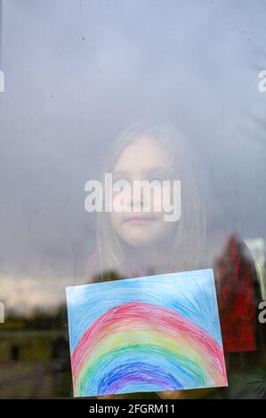 Laute Wirkung. Kind Mädchen sieben Jahre alt mit Zeichnung Regenbogen schaut durch das Fenster während covid-19 Quarantäne. Zu Hause bleiben, lassen Sie uns alle gut sein. verti Stockfoto