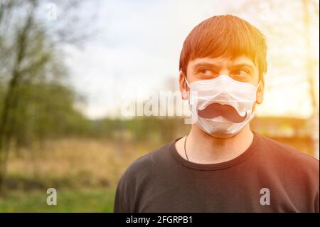 Vatertag während des covid-19 Quarantänekonzepts. Portrait eines jungen europäischen Mannes in einer Schutzmaske und einem Papierschnurrbart vor dem Hintergrund von Stockfoto