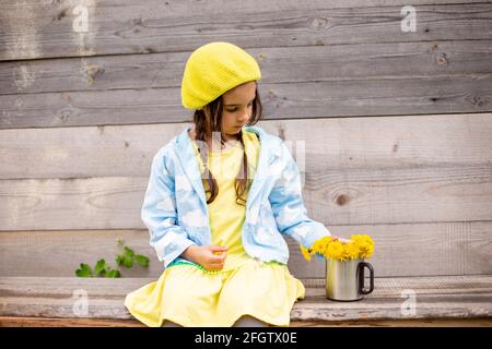 Entzückendes kleines Mädchen in einer gelben Baskenmütze, blauer Regenmantel mit Bouquet-Dandelionen in Tasse in der Nähe einer alten Holzwand Stockfoto