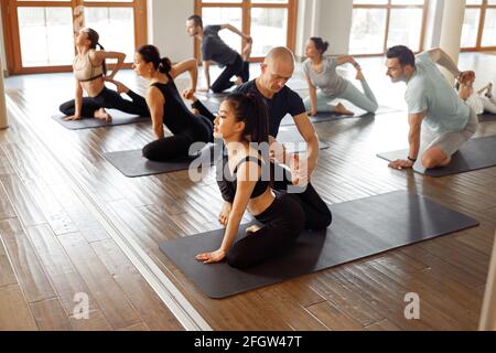 Gruppe von Menschen, die Yoga-Kurs im Fitnessclub lernen. Mann kaukasischen Ausbilder Coaching und korrigieren richtige Pose zu asiatischen Mädchen Student, während andere Stockfoto