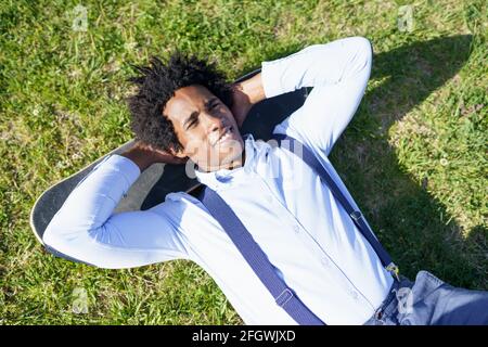 Schwarzer Arbeiter mit Afro-Haaren, die auf dem liegen Grass mit seinem Skateboard Stockfoto