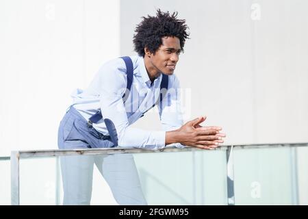 Attraktiver schwarzer Geschäftsmann mit Afro-Haaren, der sich auf einem Geländer seines Bürogebäudes ruht und den Horizont betrachtet. Stockfoto