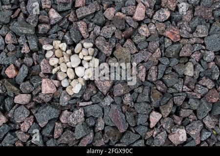 Ein Herz aus kleinen weißen, abgerundeten Steinen auf dem Hintergrund der grauen, rosa und braunen Granitsteine des Bahndamms. Konzept: Ein pur Stockfoto