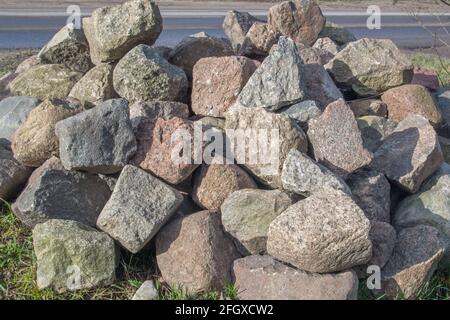 Ein Haufen Granitsteine mit scharfkantigen Kanten in Grau, Beige und Braun, die sich auf dem Gras am Straßenrand übereinander stapeln. Mineralisches Material Stockfoto
