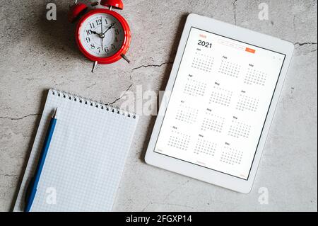 Ein Tablet mit einem offenen Kalender für 2021 Jahre, einem roten Wecker und einem Frühlingsnotizbuch mit einem Stift auf grauem Betongrund Stockfoto
