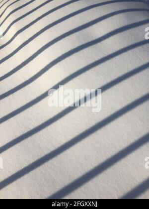 Der Schatten des Zauns auf dem Schnee an einem sonnigen Tag. Diagonale parallele Schattenlinien auf Weiß Stockfoto