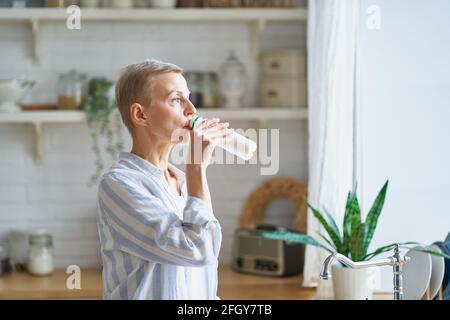 Seitenansicht einer wunderschönen älteren Frau, die Joghurt trinkt und durch das Fenster schaut, während sie zu Hause in der modernen Küche steht. Reife Menschen und gesunde Ernährung Konzept Stockfoto